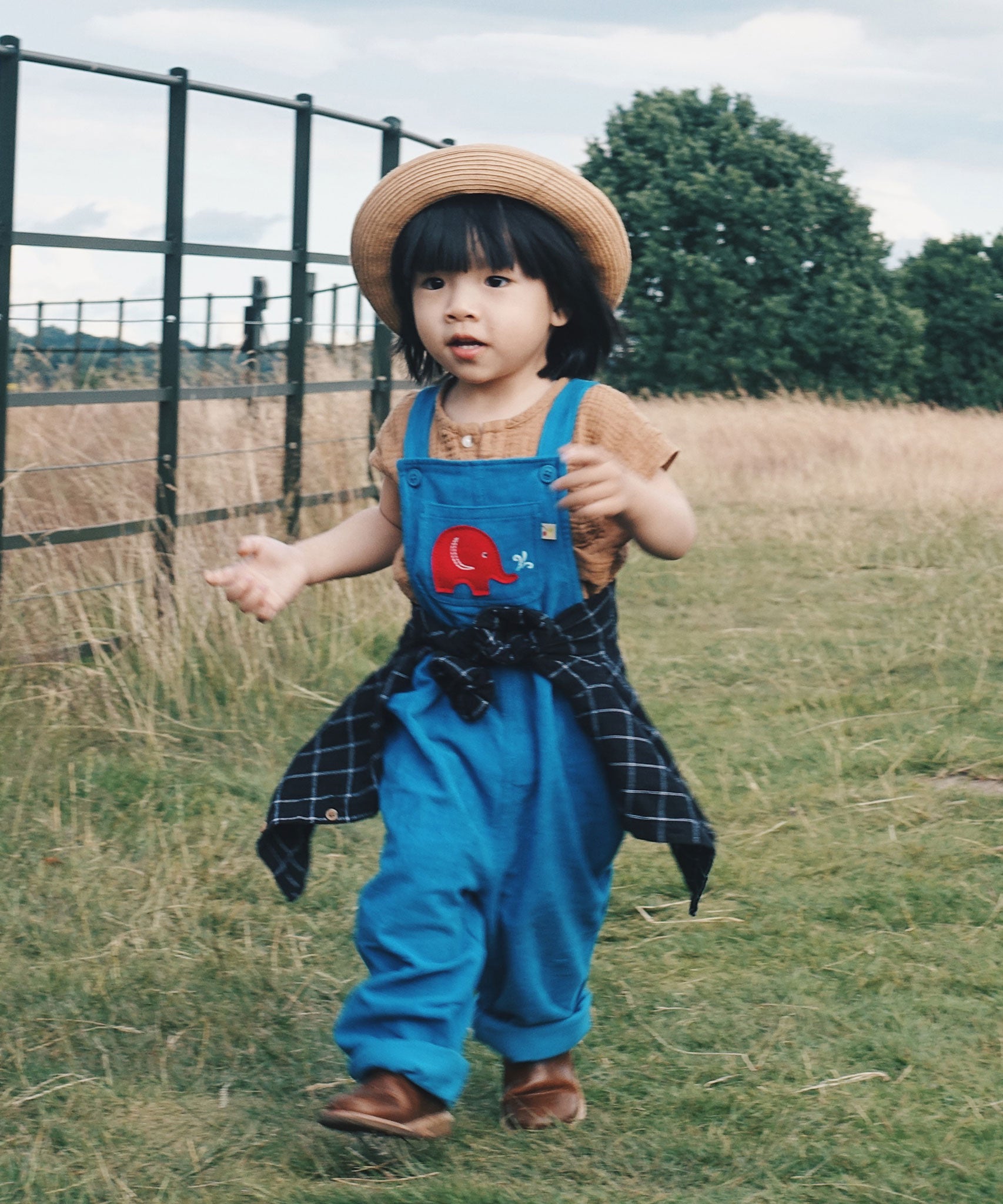 A child wearing teh blue Frugi Babipur cord dungarees with a straw hat and a black checked shirt tied around his waist. 