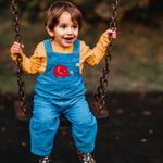 A child sitting on a swing wearing the blue Frugi Babipur cord dungarees with a yellow top underneath. 