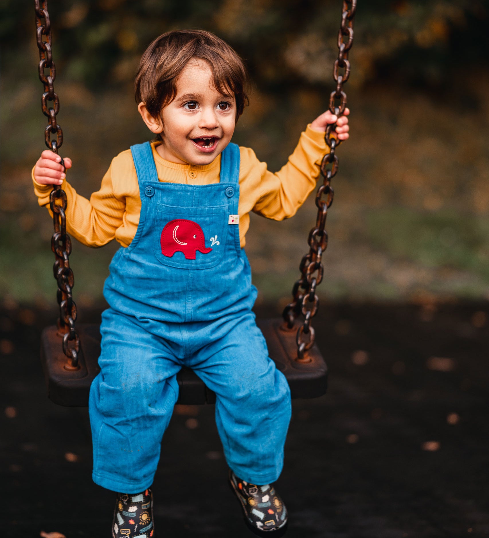 A child sitting on a swing wearing the blue Frugi Babipur cord dungarees with a yellow top underneath. 
