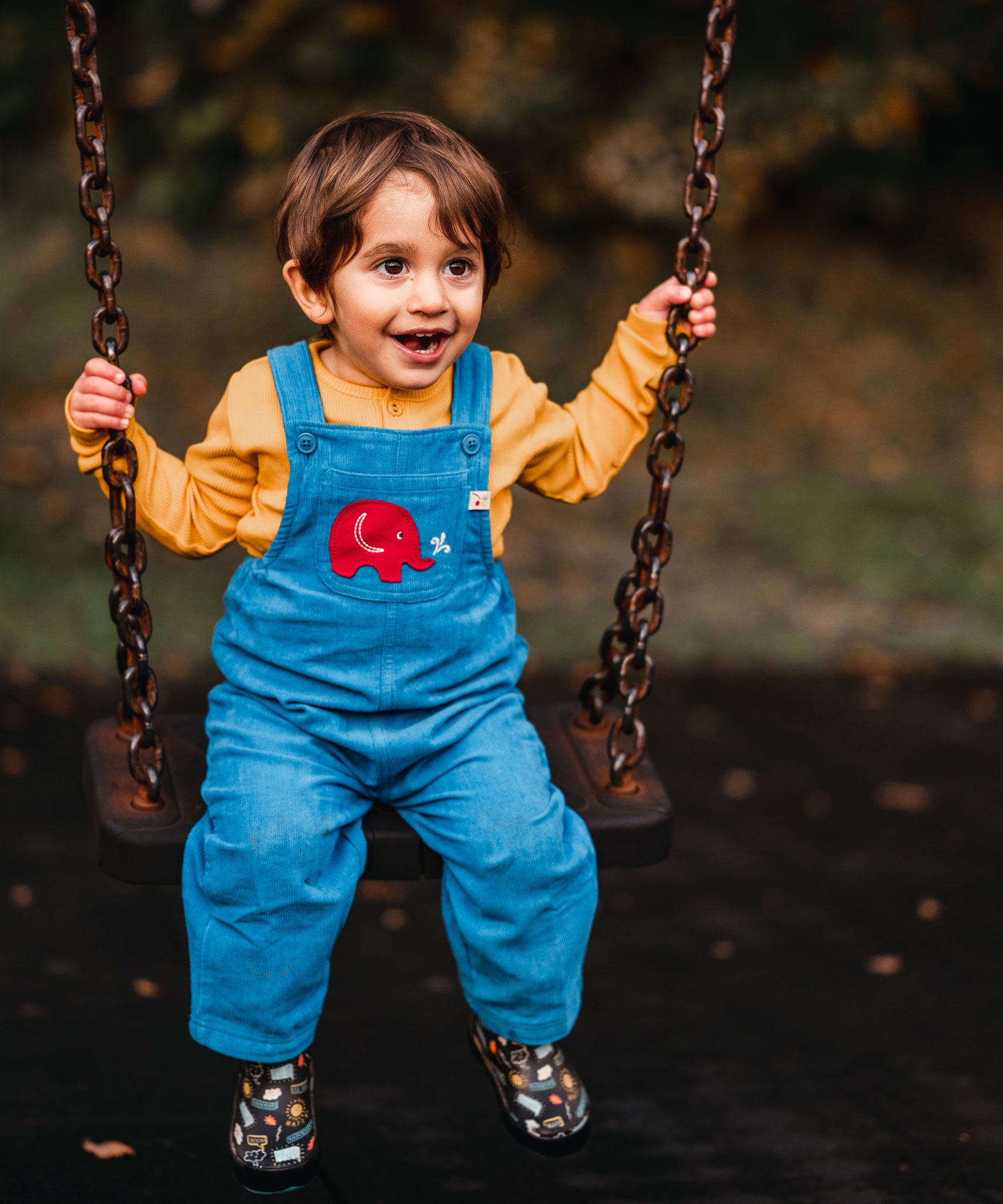 A child sitting on a swing wearing the blue Frugi Babipur cord dungarees with a yellow top underneath. 