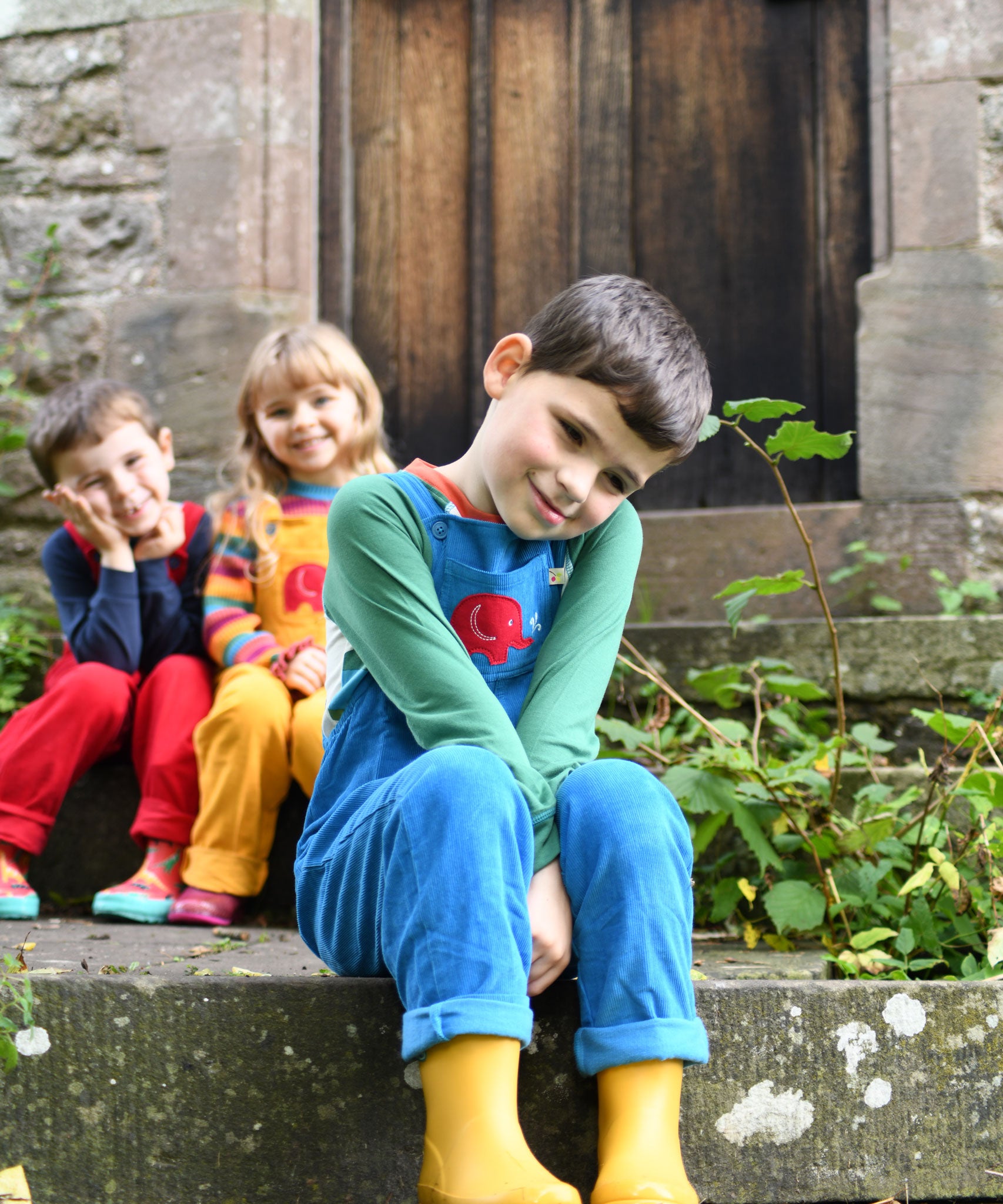 A child sitting on  steps outdoors wearing the blue Frugi Babipur cord dungarees. 