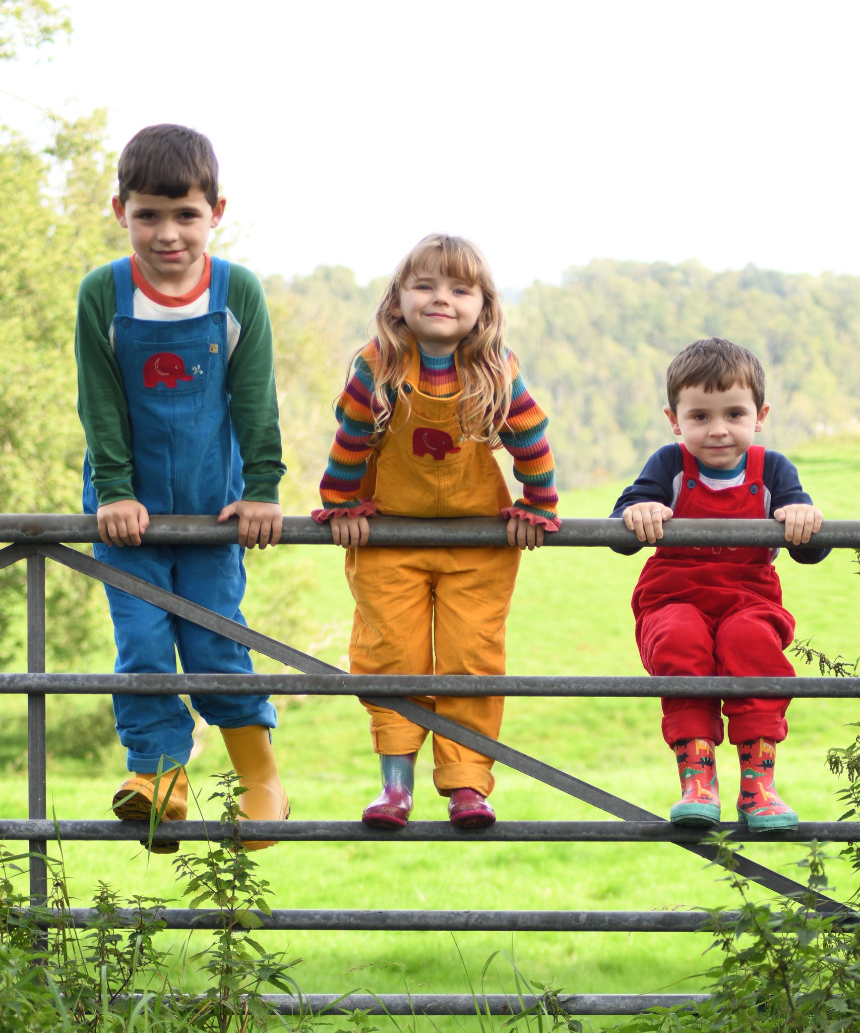 Three siblings standing on a metal gate. They are all wearing Frugi Babipur cord dungarees,  one in blue, one in yellow and one in red. 