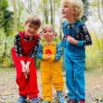 Three siblings standing in a forest. They are all wearing Frugi Babipur cord dungarees,  one in red, one in yellow and one in blue. 