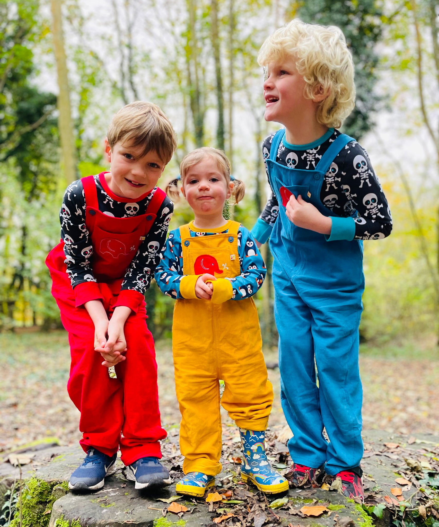 Three siblings standing in a forest. They are all wearing Frugi Babipur cord dungarees,  one in red, one in yellow and one in blue. 