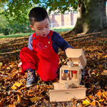 A child wearing the red cord Frugi Babipur dungarees . The child is kneeling on a leaf covered floor under a tree playing with a wooden Fagus truck. 