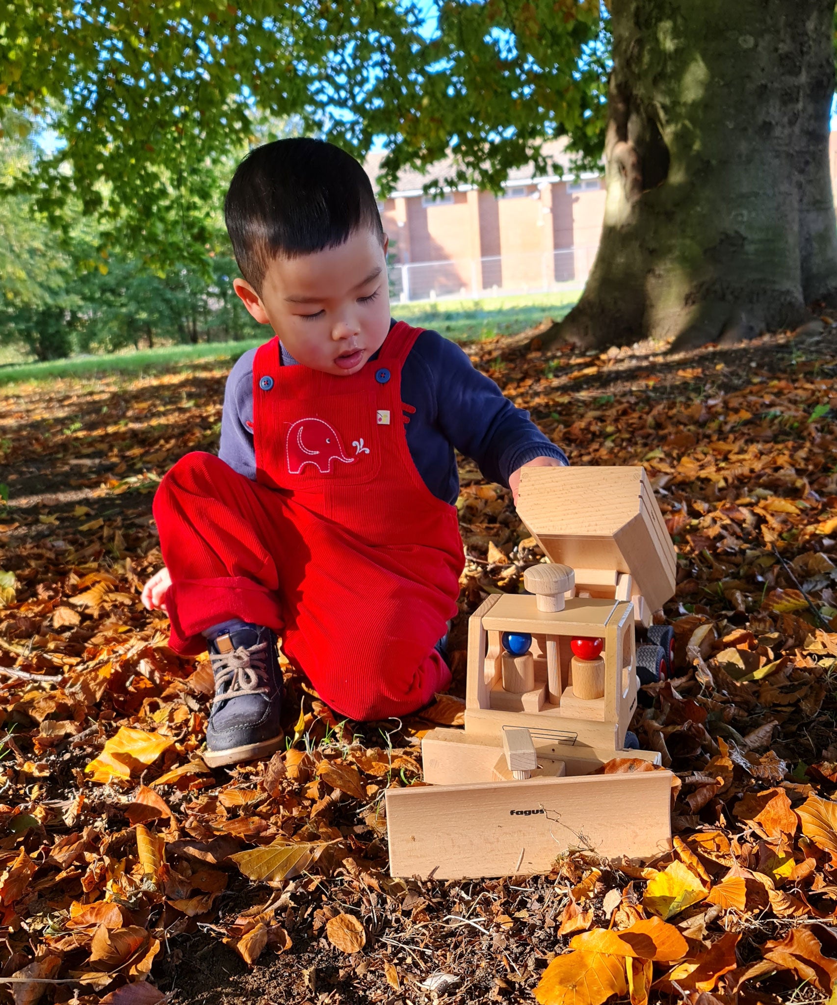 A child wearing the red cord Frugi Babipur dungarees . The child is kneeling on a leaf covered floor under a tree playing with a wooden Fagus truck. 