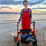 A child stood on a beach using a mobility aid wearing the red coloured Frugi Babipur cord dungarees. 