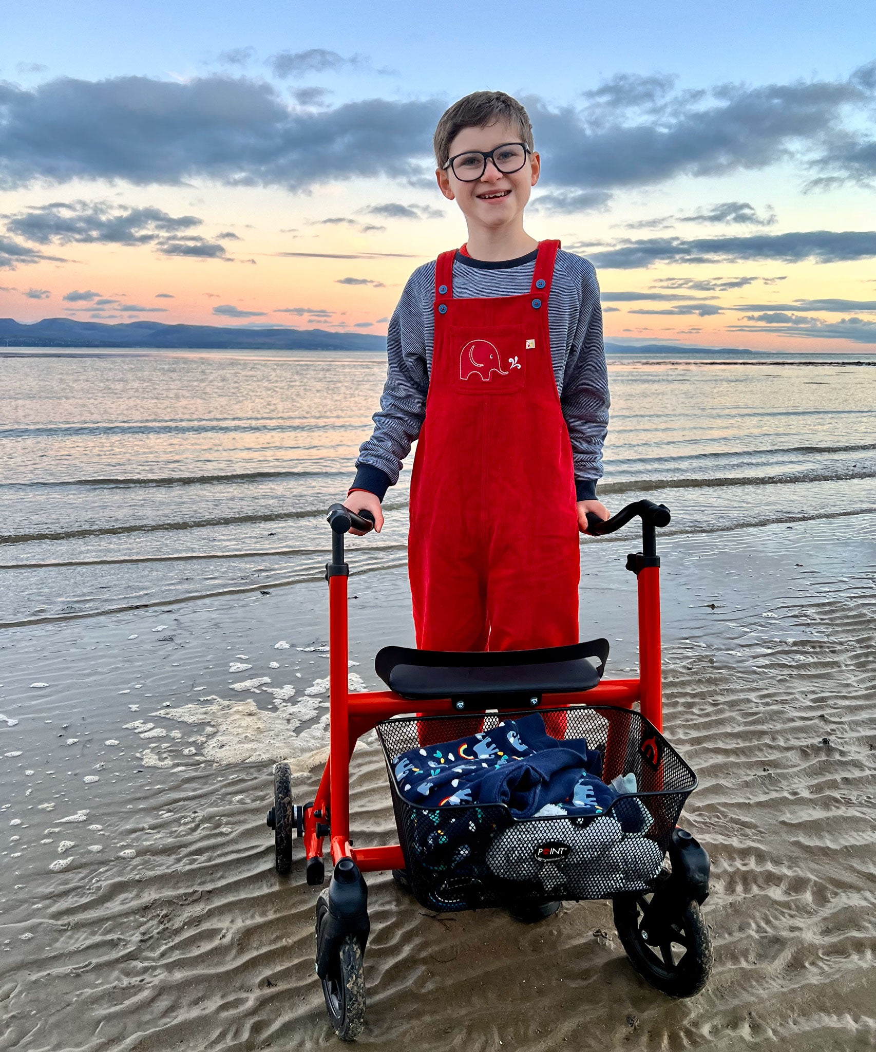 A child stood on a beach using a mobility aid wearing the red coloured Frugi Babipur cord dungarees. 