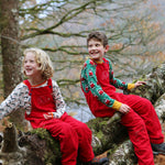 Two children sitting on a moss covered tree branch, both are wearing the red coloured Frugi Babipur cord dungarees, 