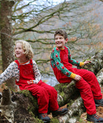 Two children sitting on a moss covered tree branch, both are wearing the red coloured Frugi Babipur cord dungarees, 