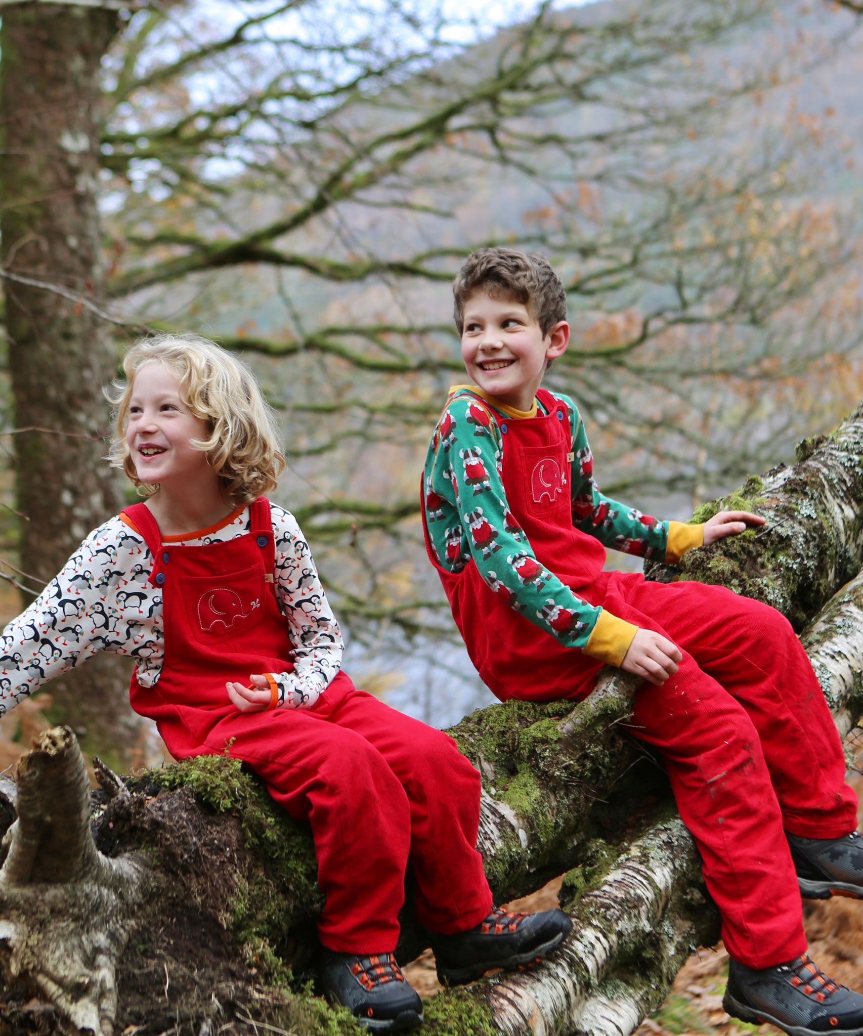 Two children sitting on a moss covered tree branch, both are wearing the red coloured Frugi Babipur cord dungarees, 