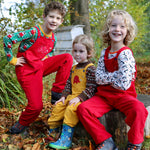 Three children sitting on a large tree stump. Two are wearing the red Frugi Babipur cord dungarees and one is wearing teh yellow version. 
