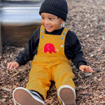 A child sitting on bark on teh floor of a playground n front of a metal slide. The child is wearing a yellow Frugi Babipur cord dungarees.