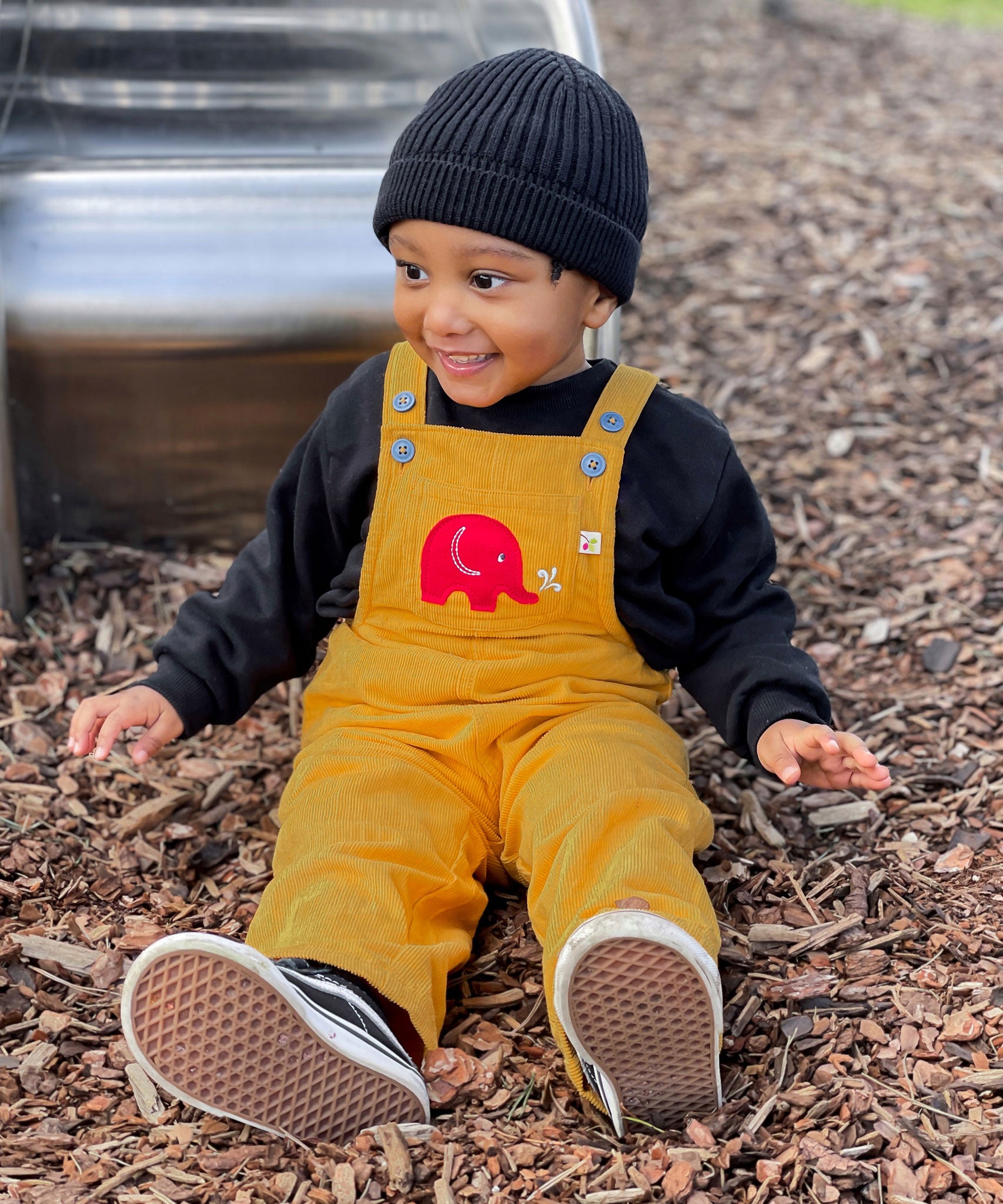 A child sitting on bark on teh floor of a playground n front of a metal slide. The child is wearing a yellow Frugi Babipur cord dungarees.