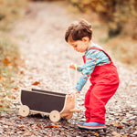 A child wearing a red Frugi Babipur cord dungarees pulling along a Plan Toys wagon on a leaf covered path. The side of teh dungarees can be seen. 