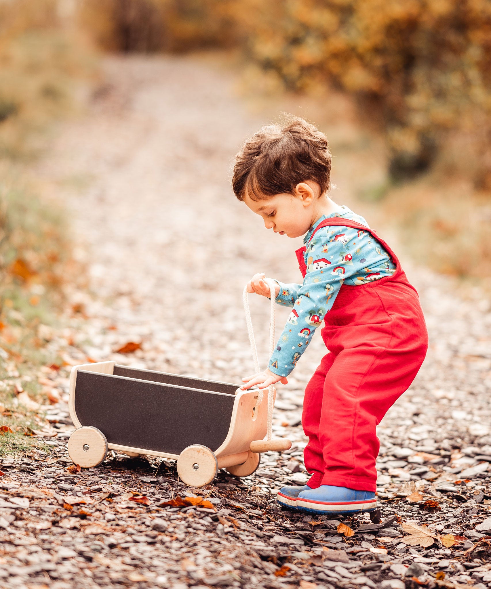 A child wearing a red Frugi Babipur cord dungarees pulling along a Plan Toys wagon on a leaf covered path. The side of teh dungarees can be seen. 