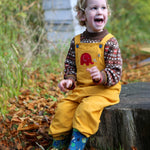 A child sitting on a large tree stump smiling and wearing a Frugi Babipur yellow cord dungarees. 