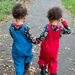 Twins wearing the Frugi Babipur dungarees, one in red, one in blue. They are walking away from the camera showing the back of the dungarees. 