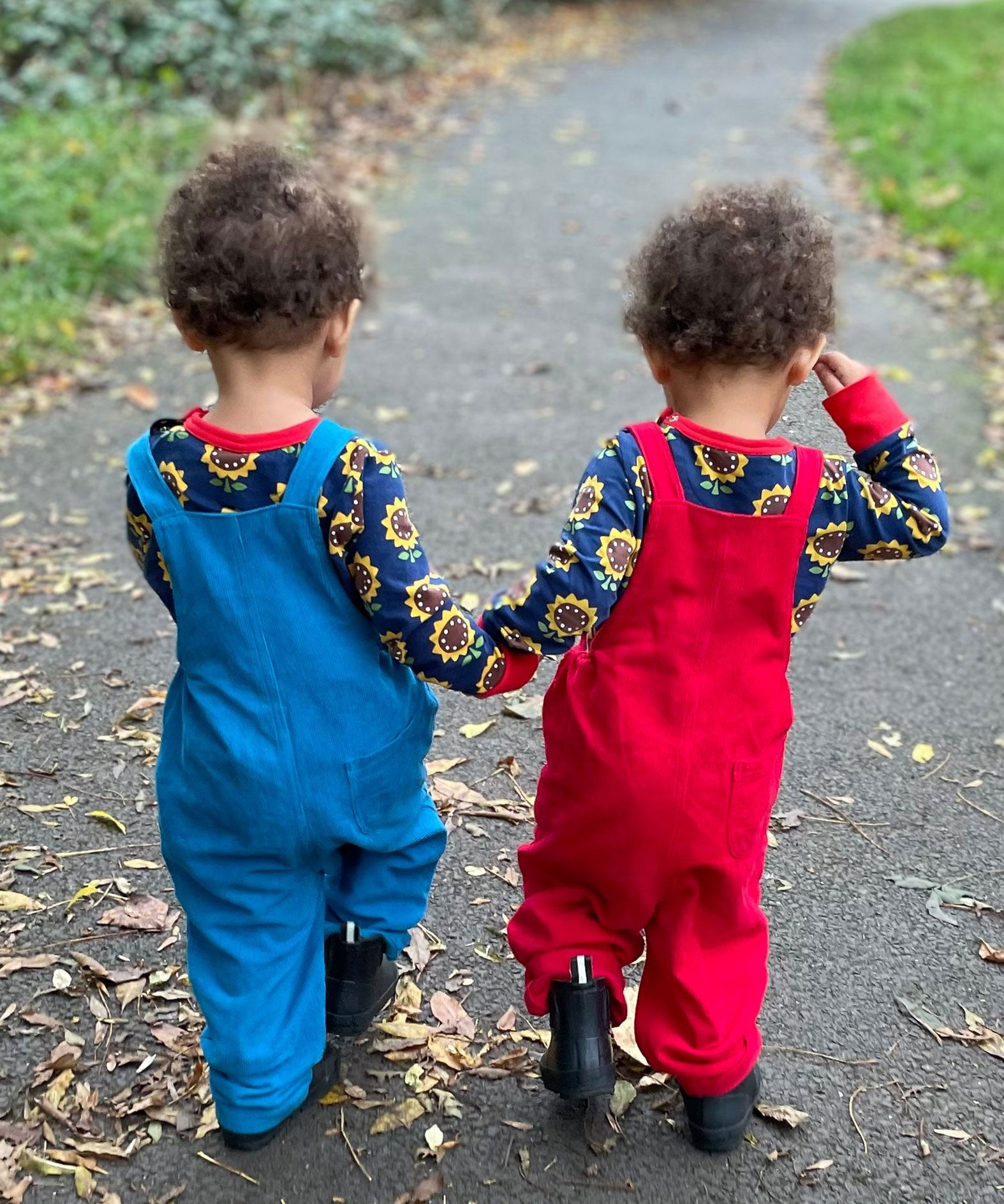 Twins wearing the Frugi Babipur dungarees, one in red, one in blue. They are walking away from the camera showing the back of the dungarees. 