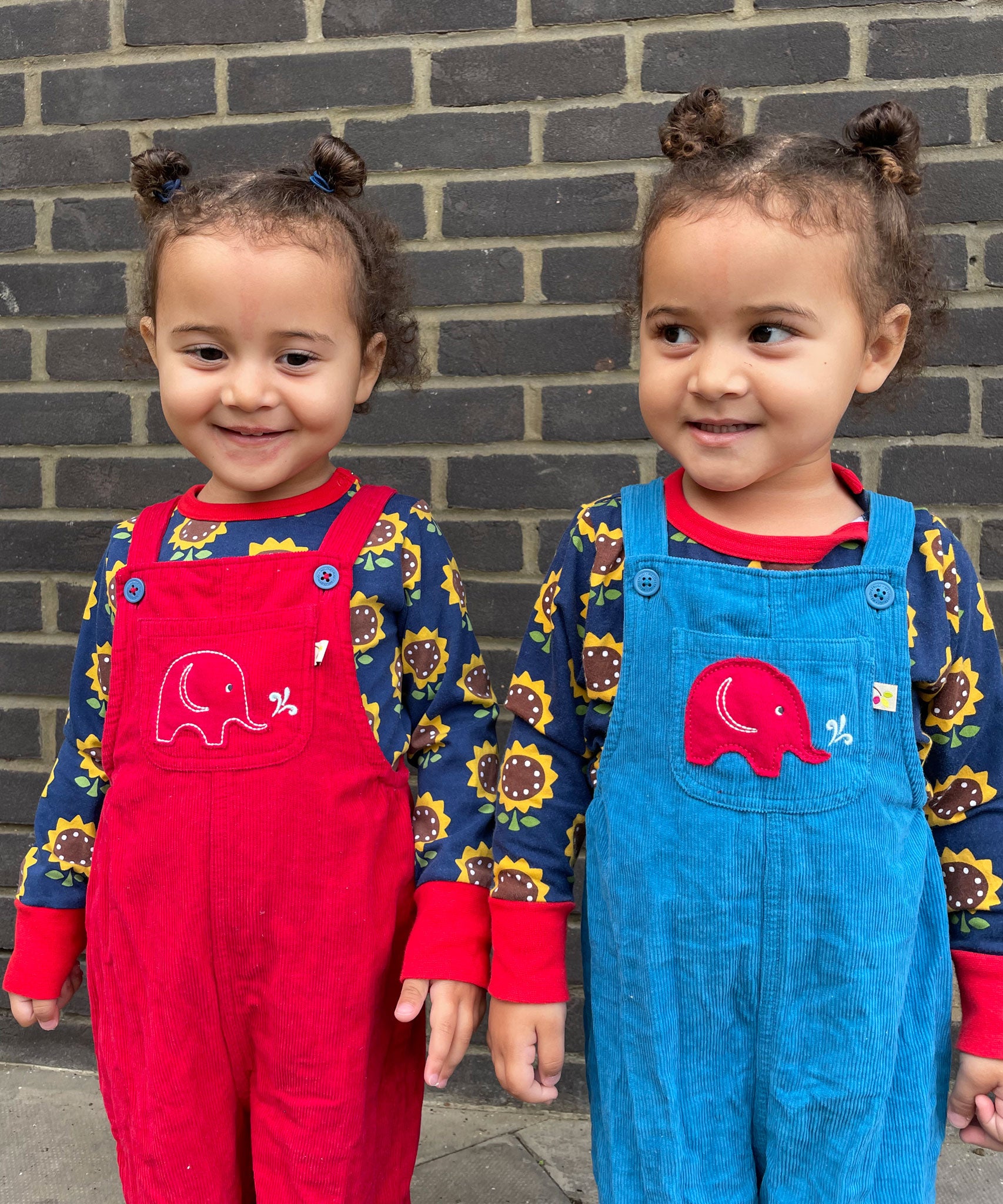 Twins wearing the Frugi Babipur dungarees, one in red, one in blue. 