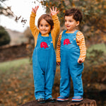 Two children standing on a tree stump wearing the blue Frugi Babipur cord dungarees. Both children are wearing yellow long sleeved tops underneath, one is holding both their hands up in the air. 