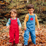 Two children smiling playing in a forest standing on a leaf covered floor. One is wearing the red Frugi Babipur cord dungarees with teh other wearing the blue ones. 