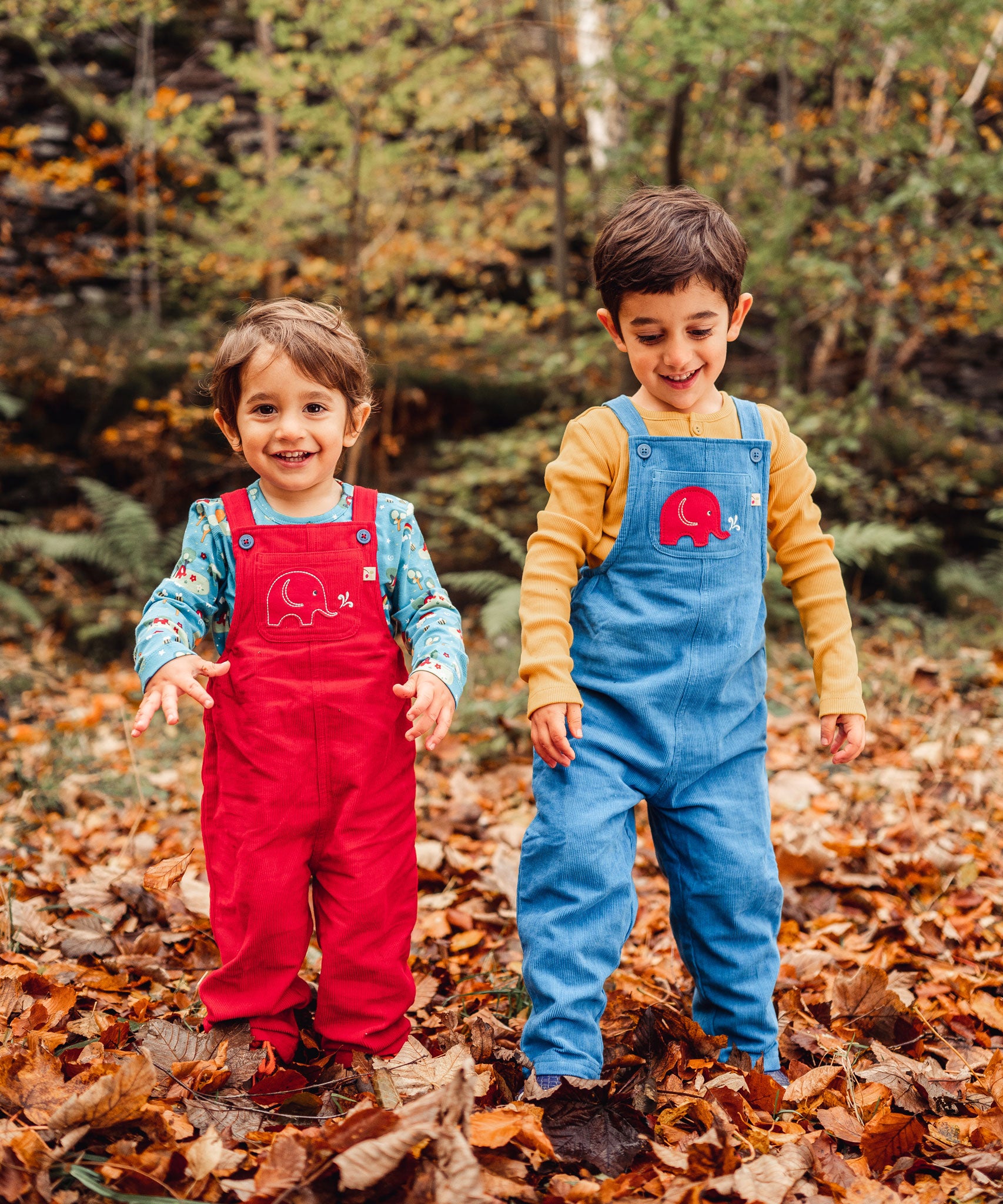 Two children smiling playing in a forest standing on a leaf covered floor. One is wearing the red Frugi Babipur cord dungarees with teh other wearing the blue ones. 