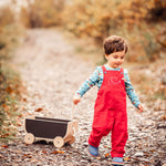 A child wearing a red Frugi Babipur cord dungarees pulling along a Plan Toys wagon on a leaf covered path. 