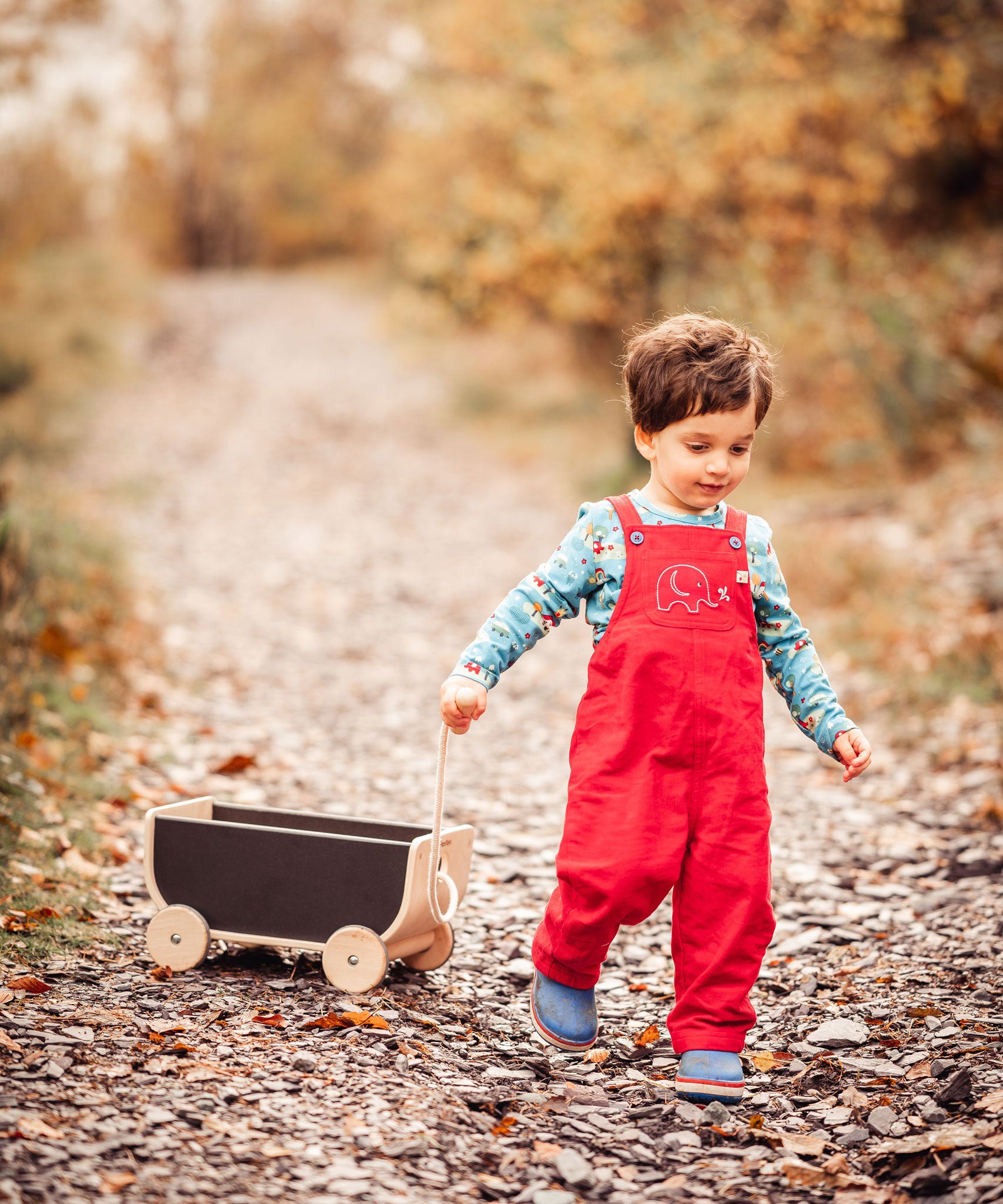 A child wearing a red Frugi Babipur cord dungarees pulling along a Plan Toys wagon on a leaf covered path. 