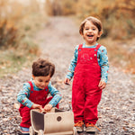 Two children outdoors playing with a Plan Toys wagon. Both are wearing red coloured Frugi Babipur corduroy dungarees.