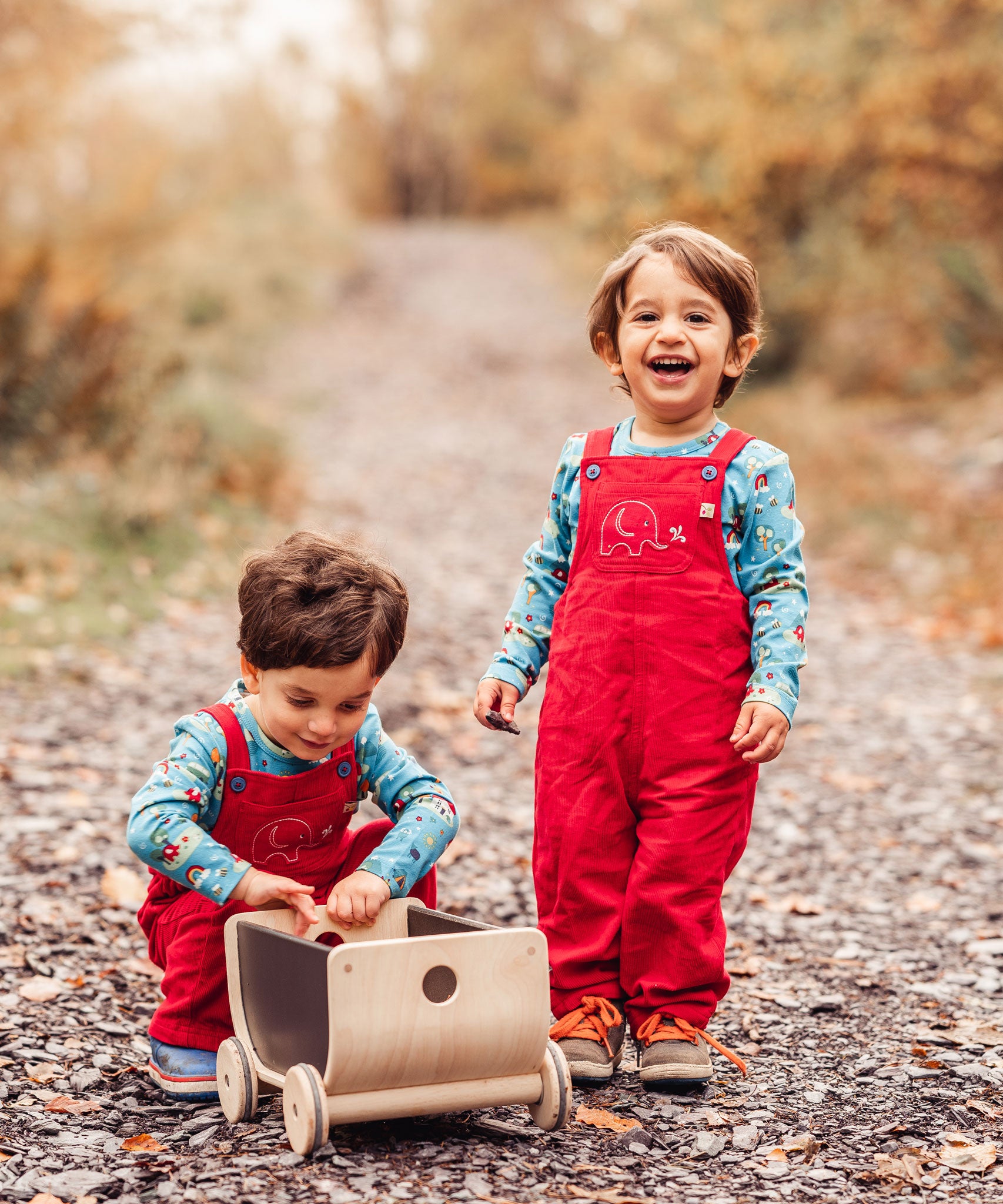 Two children outdoors playing with a Plan Toys wagon. Both are wearing red coloured Frugi Babipur corduroy dungarees.