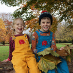 Two children sitting on a large log in park. One is wearing a yellow Frugi Babipur dungarees and the other is wearing the blue version. 