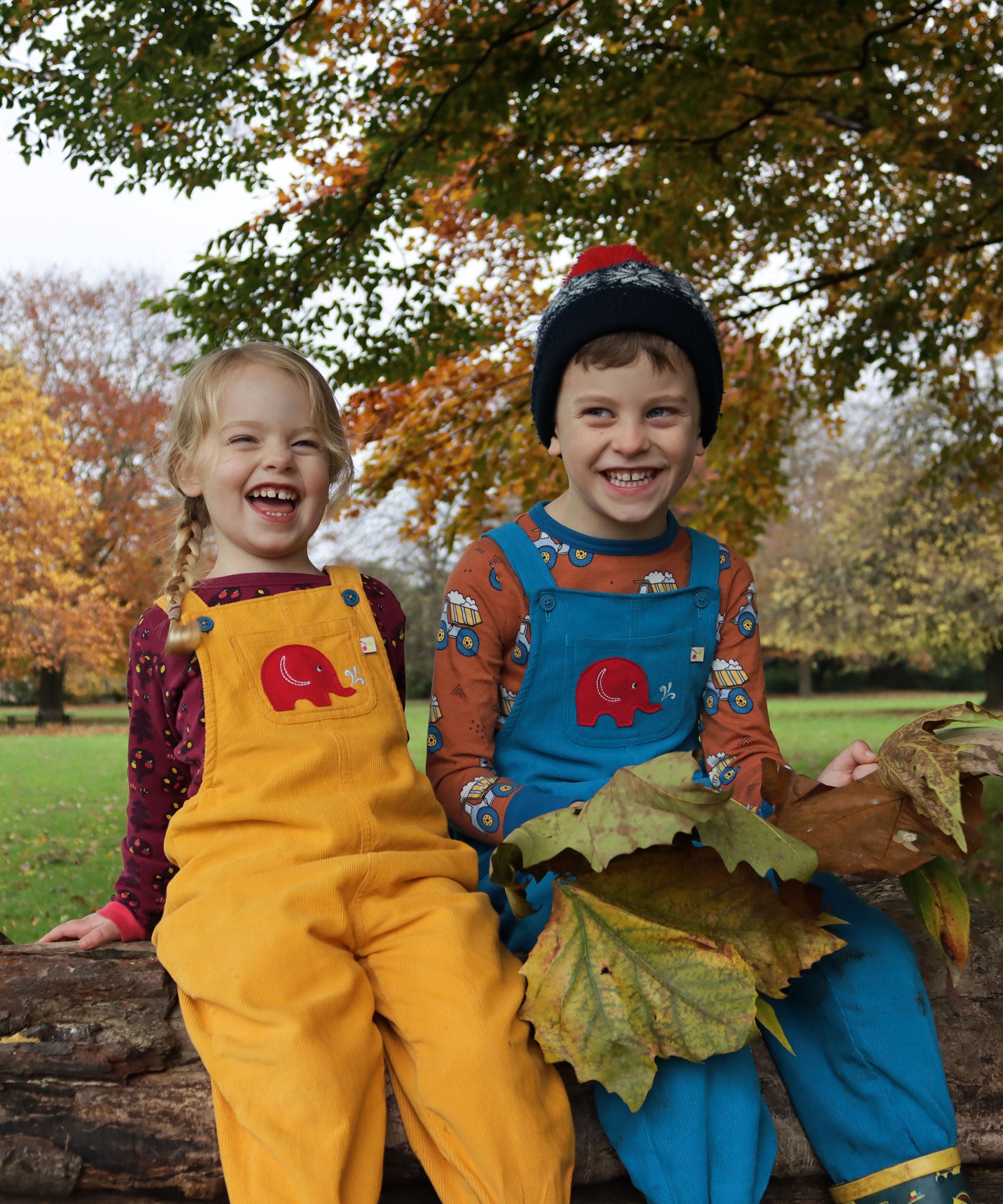 Two children sitting on a large log in park. One is wearing a yellow Frugi Babipur dungarees and the other is wearing the blue version. 