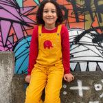 A child wearing the yellow cord Frugi Babipur dungarees sitting in front of a graffitied wall.  