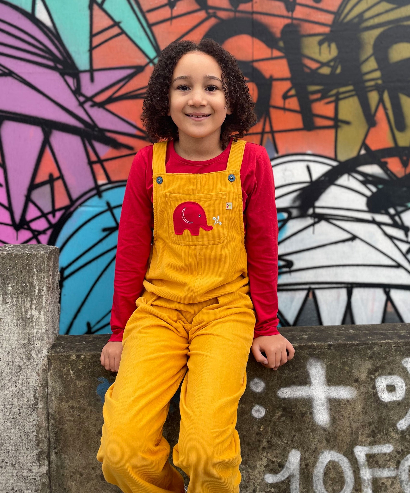 A child wearing the yellow cord Frugi Babipur dungarees sitting in front of a graffitied wall.  