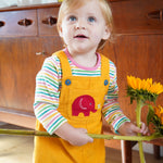 A toddler wearing the yellow cord Frugi Babipur dungarees holding onto large sunflowers. 