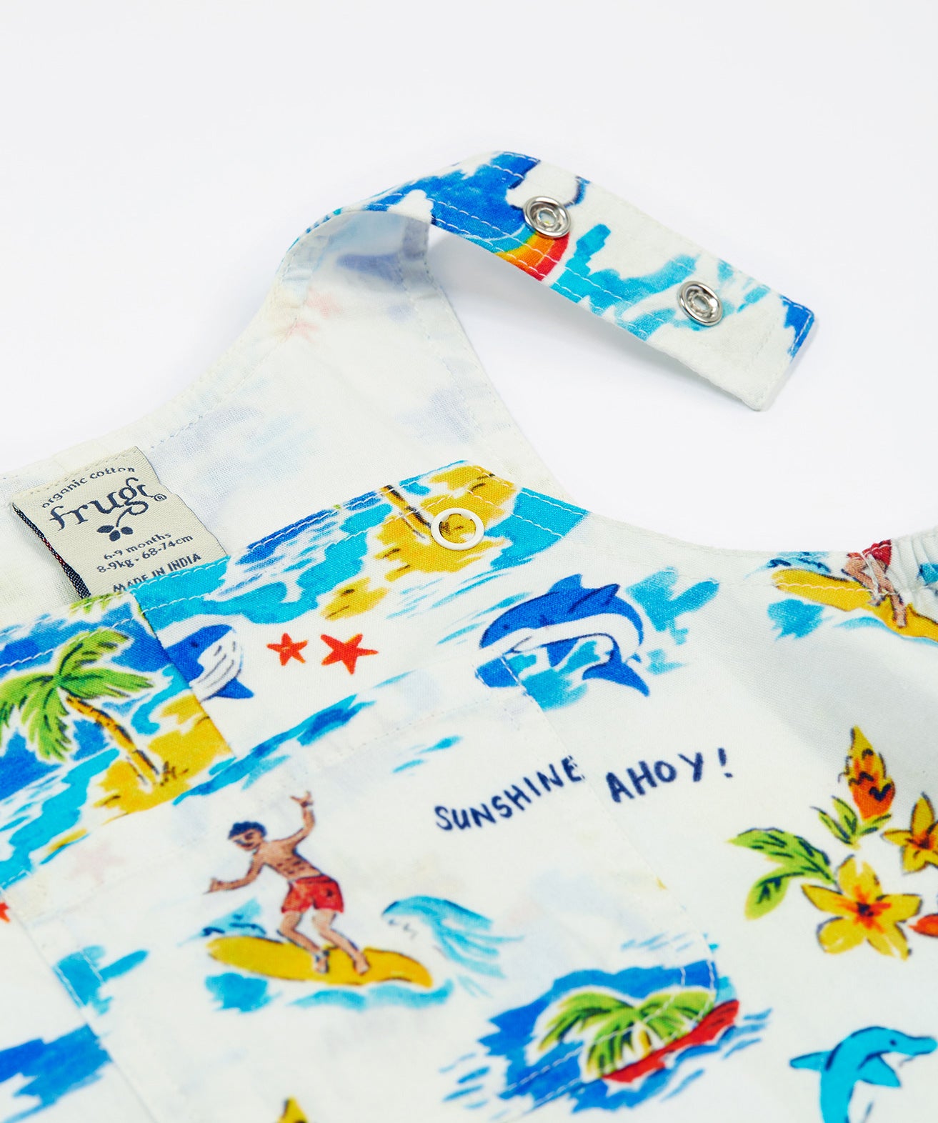 A close-up of the Frugi Aloha Organic Cotton Dungarees front pocket and popper fastenings, featuring a colourful tropical beach print with dolphins, rainbows and sailing boats.