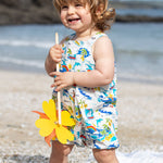 Smiling toddler wearing Frugi Aloha Organic Cotton Dungarees, featuring a colourful tropical beach print with rainbows, dolphins, and sailing boats, standing barefoot on a pebbled beach and holding a yellow pinwheel.  