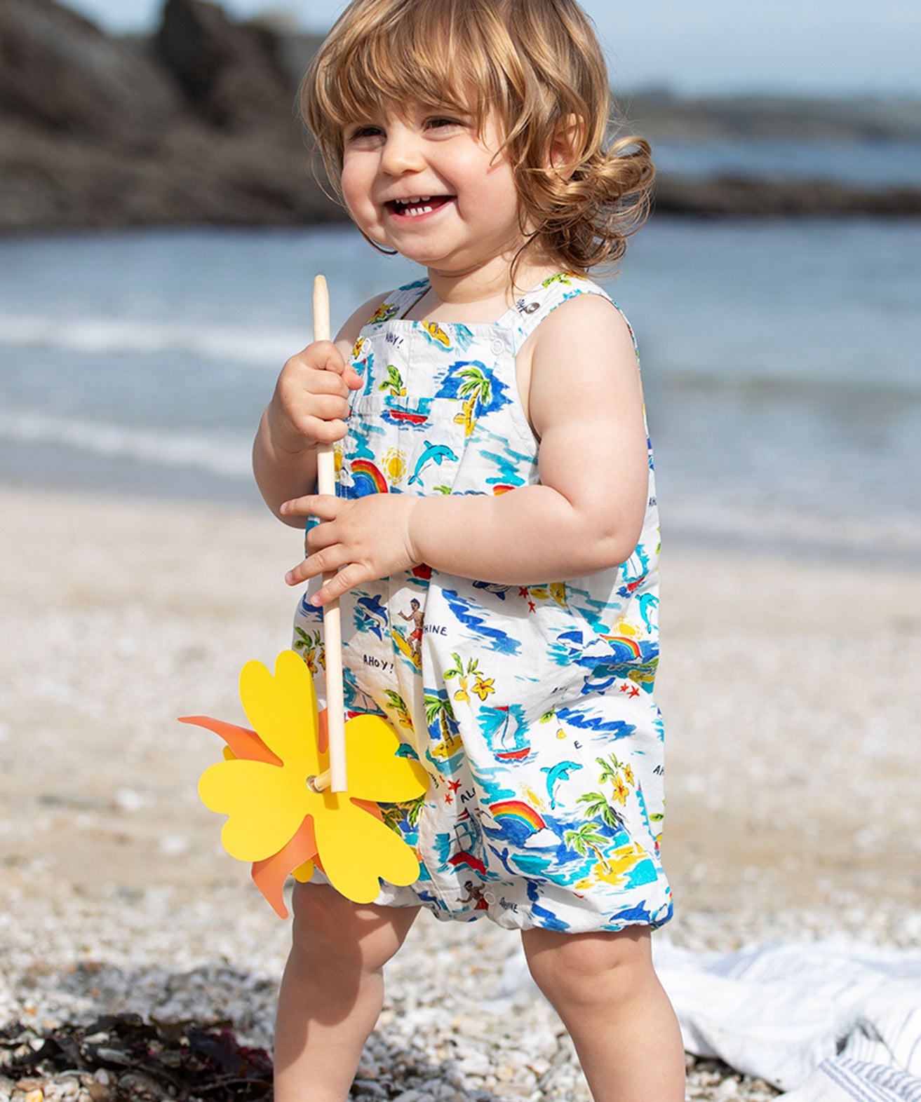 Smiling toddler wearing Frugi Aloha Organic Cotton Dungarees, featuring a colourful tropical beach print with rainbows, dolphins, and sailing boats, standing barefoot on a pebbled beach and holding a yellow pinwheel.  