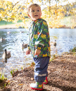 A toddler standing by a lake and wearing the Frugi puddle buster navy trousers, be hoppy coat and matching puddle buster wellington boots  A natural rubber and waterproof wellington boot in navy with red trim with prints of multicoloured frogs.