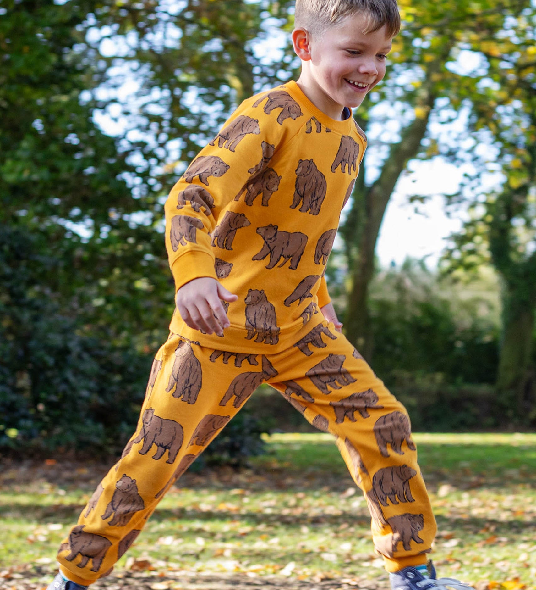 A child running outside and wearing the Frugi beary wild parsnip joggers. A dark yellow GOTS organic joggers with a brown bear print. 