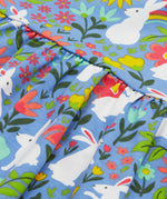 Close up of the Frugi kids Bella dress bunny bounce. Showing a collection of white rabbits printed with different coloured flowers and bright rainbows on light blue organic cotton fabric