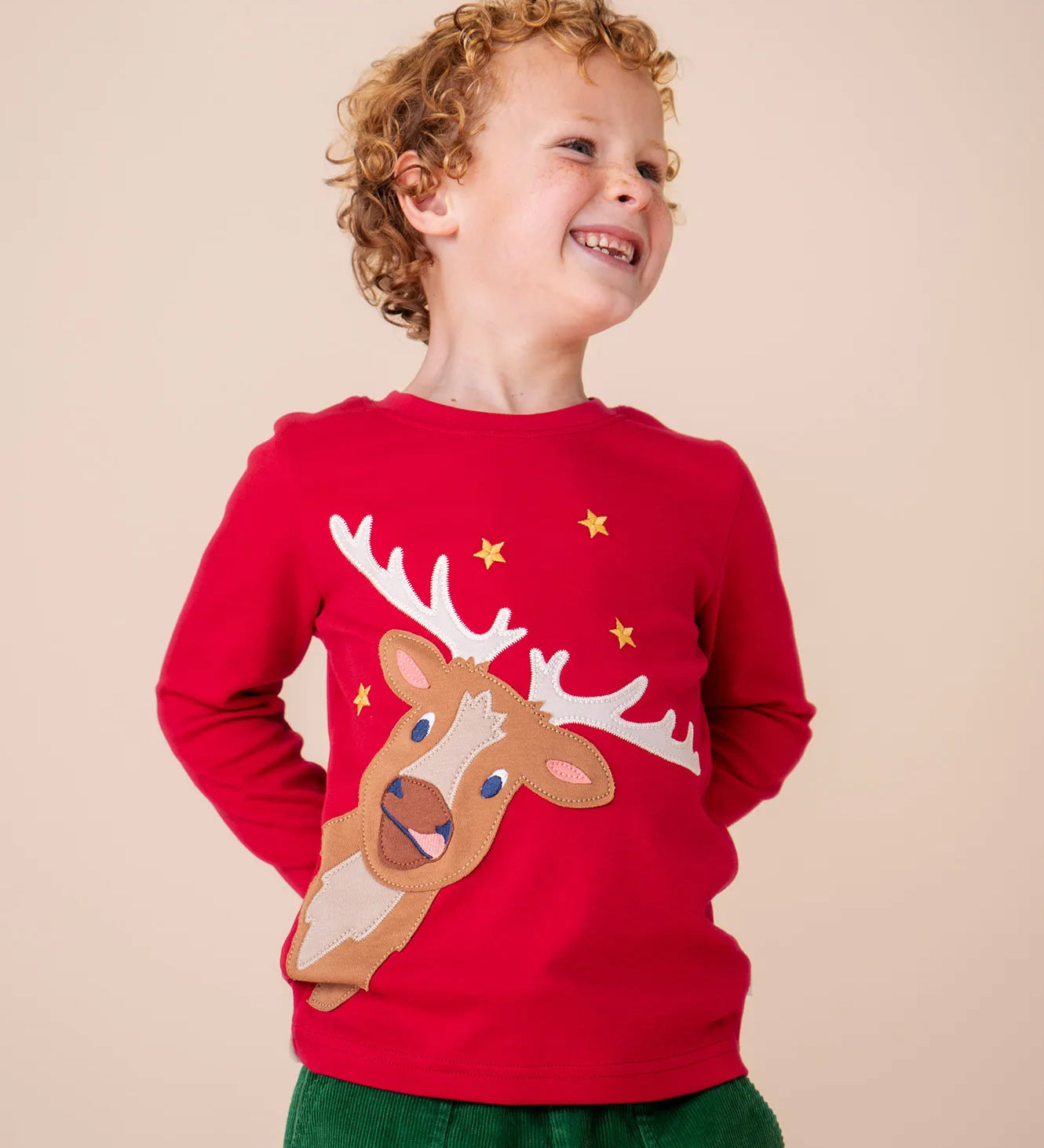 A child wearing the Frugi Berry Red Reindeer Terrific Circular Organic Cotton long sleeved kids Top available at Babipur. A red coloured children's top with a festive reindeer applique and gold stars on the front. 