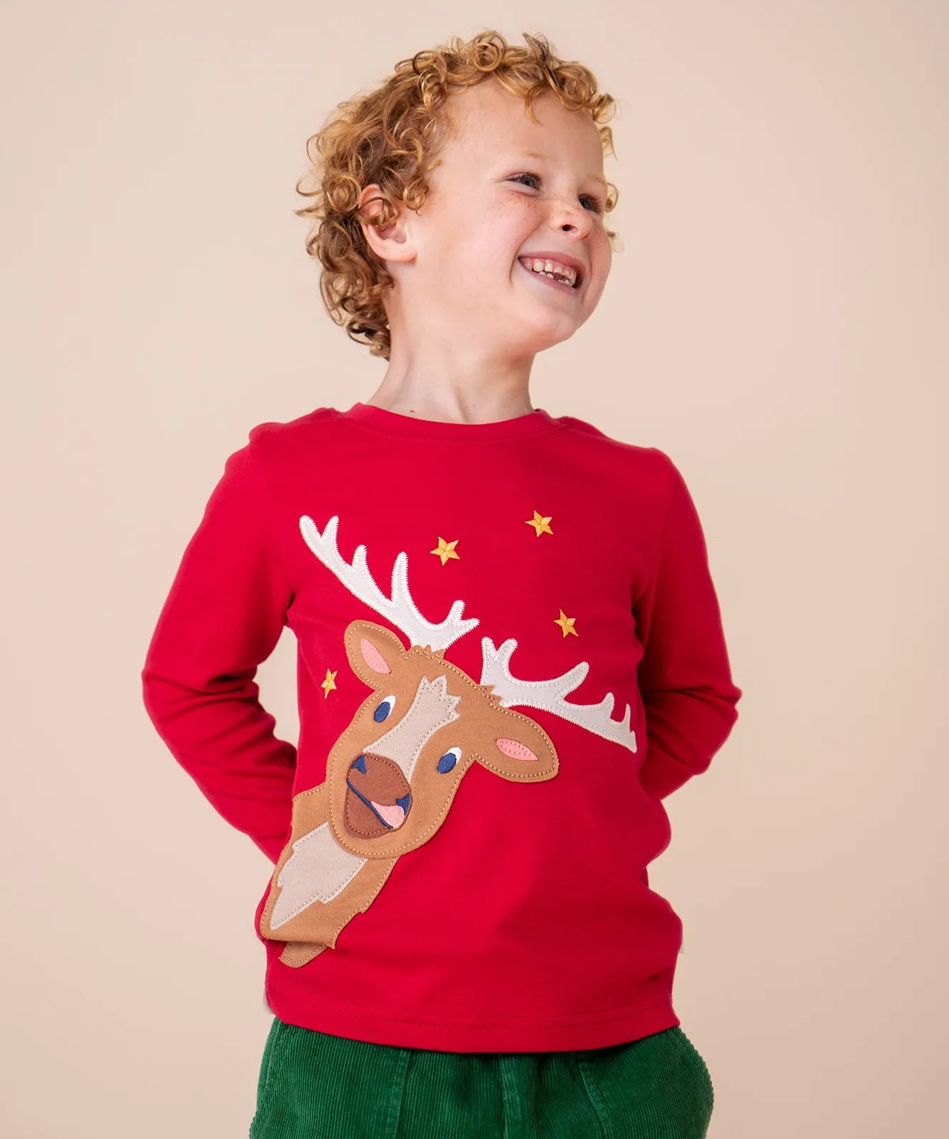 A child wearing the Frugi Berry Red Reindeer Terrific Circular Organic Cotton long sleeved kids Top available at Babipur. A red coloured children's top with a festive reindeer applique and gold stars on the front. 