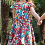 A young child wearing Frugi Morwenna Skater Dress in the Birds Of Paradise print. Featuring different exotic birds surrounded by colourful flowers and leaves on a pink background, walking holding hands amongst palm trees.