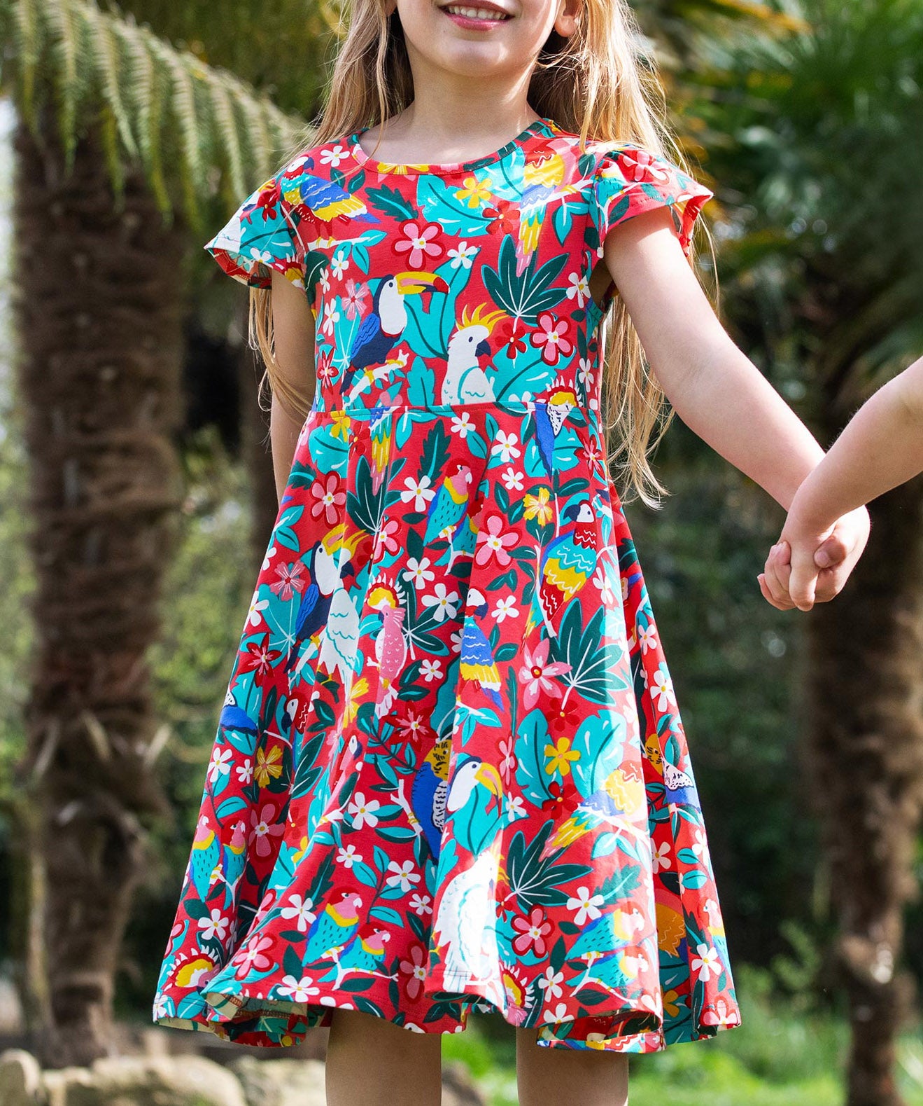 A young child wearing Frugi Morwenna Skater Dress in the Birds Of Paradise print. Featuring different exotic birds surrounded by colourful flowers and leaves on a pink background, walking holding hands amongst palm trees.