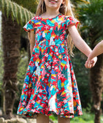 A young child wearing Frugi Morwenna Skater Dress in the Birds Of Paradise print. Featuring different exotic birds surrounded by colourful flowers and leaves on a pink background, walking holding hands amongst palm trees.
