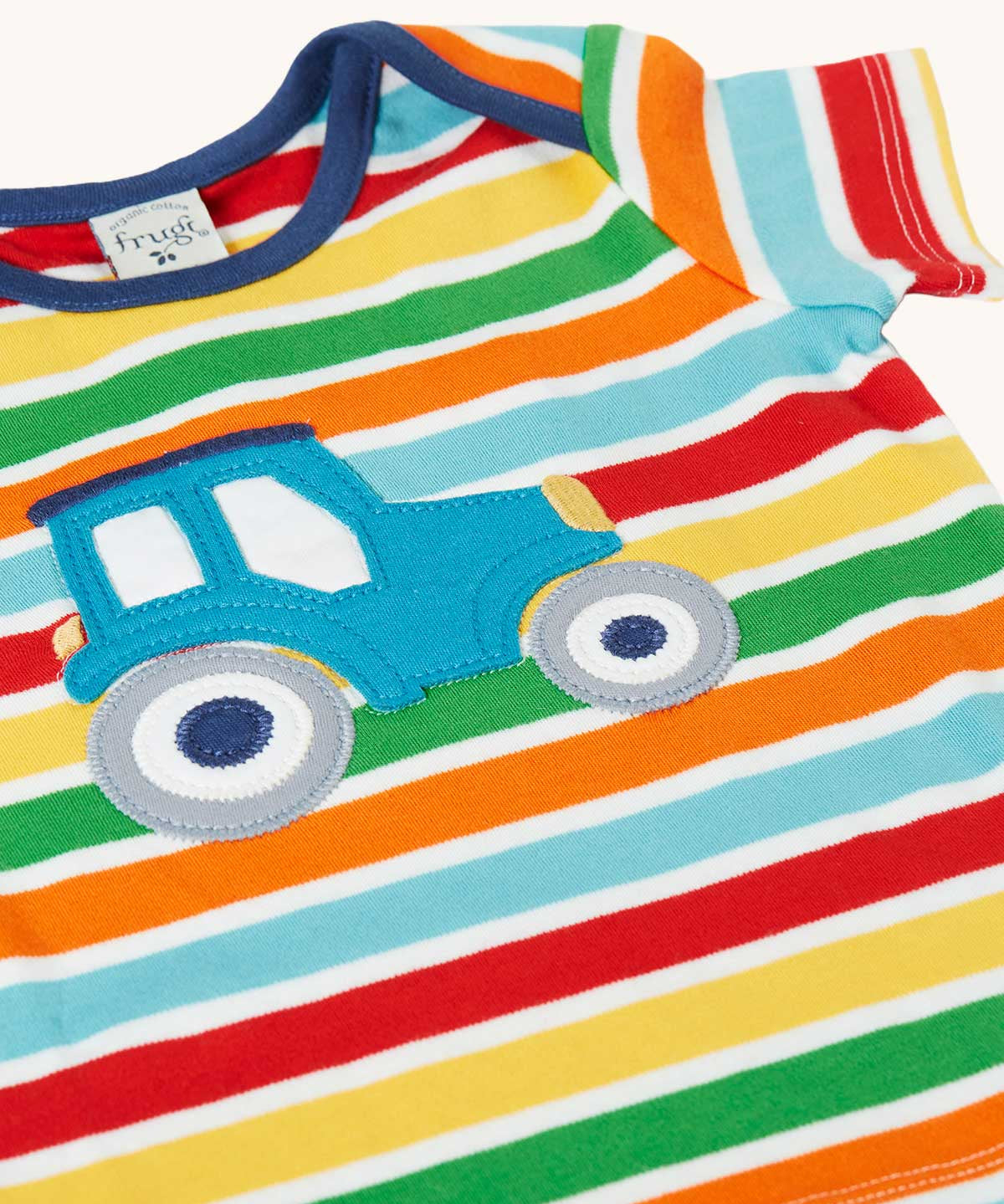 Close up of the front of the Frugi blue tractor applique rainbow striped t-shirt. Bands of red, light yellow, green, orange and light blue in-between thinner white stripes.