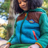 A child sitting on a tree and wearing the Frugi kids braken fleece cord jacket in holly green/hotchpotch.