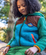 A child sitting on a tree and wearing the Frugi kids braken fleece cord jacket in holly green/hotchpotch.